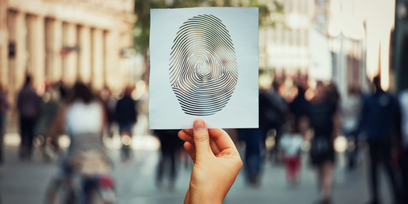 A hand holding a white sheet with a large fingerprint illustration against a blurred city street background, symbolising identity verification for UK company directors and PSCs under Companies House 2025 compliance rules.